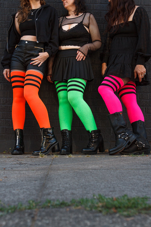 3 people wearing Electric Shock thigh highs in neon green, neon orange, and neon pink, all with black stripes.