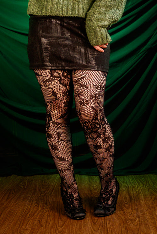 Person wearing a green sweater, black patterned floral tights, and a black skirt against a green curtain.