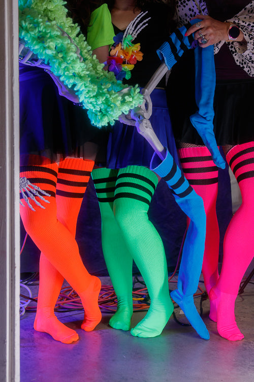 3 people and one plastic skeleton wearing Electric Shock thigh highs in neon blue, neon green, neon orange, and neon pink, all with black stripes.