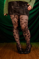 Person wearing a green sweater, black patterned floral tights, and a black skirt against a green curtain.